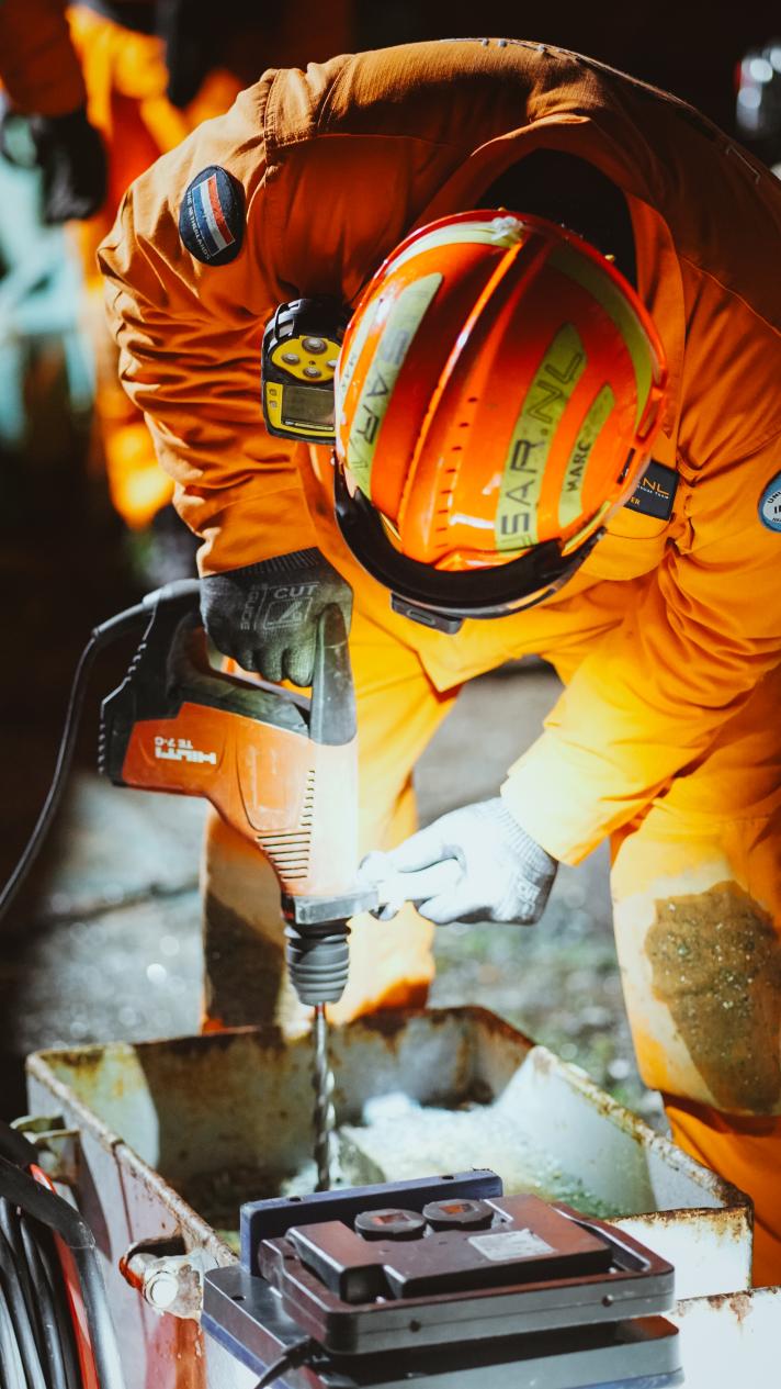 A man wearing bright orange protective clothing and helmet drills into a concrete slab.