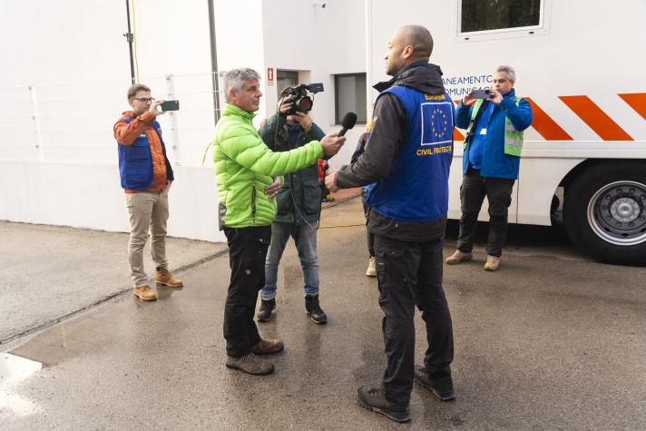 A man wearing a UCPM jacket speaks into a microphone. A video crew film the interview.